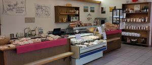 Eine Bäckerei mit Holzregalen, gefüllt mit Brot und Gebäck. Ein Ausstellungsfall mit verschiedenen Desserts steht auf dem Tresen, und ein weiteres Regal hält mehr Brot. Verschiedene Gegenstände hängen an der Wand.