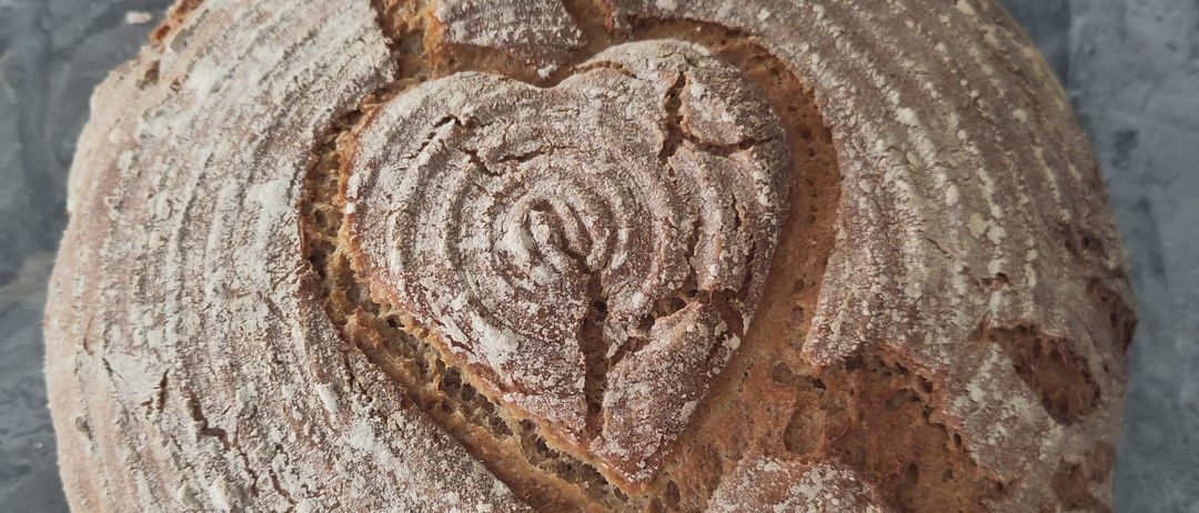 Ein runder Laib rustikales Brot mit einem herzförmigen Schnitt auf der Oberseite liegt auf einer Marmoroberfläche. Das Brot hat eine goldbraune Kruste und eine leicht rissige Oberfläche.