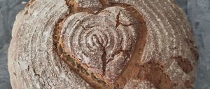 Ein runder Laib rustikales Brot mit einem herzförmigen Schnitt auf der Oberseite liegt auf einer Marmoroberfläche. Das Brot hat eine goldbraune Kruste und eine leicht rissige Oberfläche.