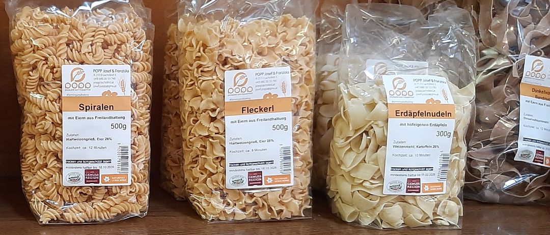 Ein Holzregal mit mehreren Tüten Pasta. Von links nach rechts: Bandnudeln, Spiralen, Elenkerl und Erdäpfelnudeln. Alle Tüten haben weiße Etiketten mit Text und Nährwertangaben.