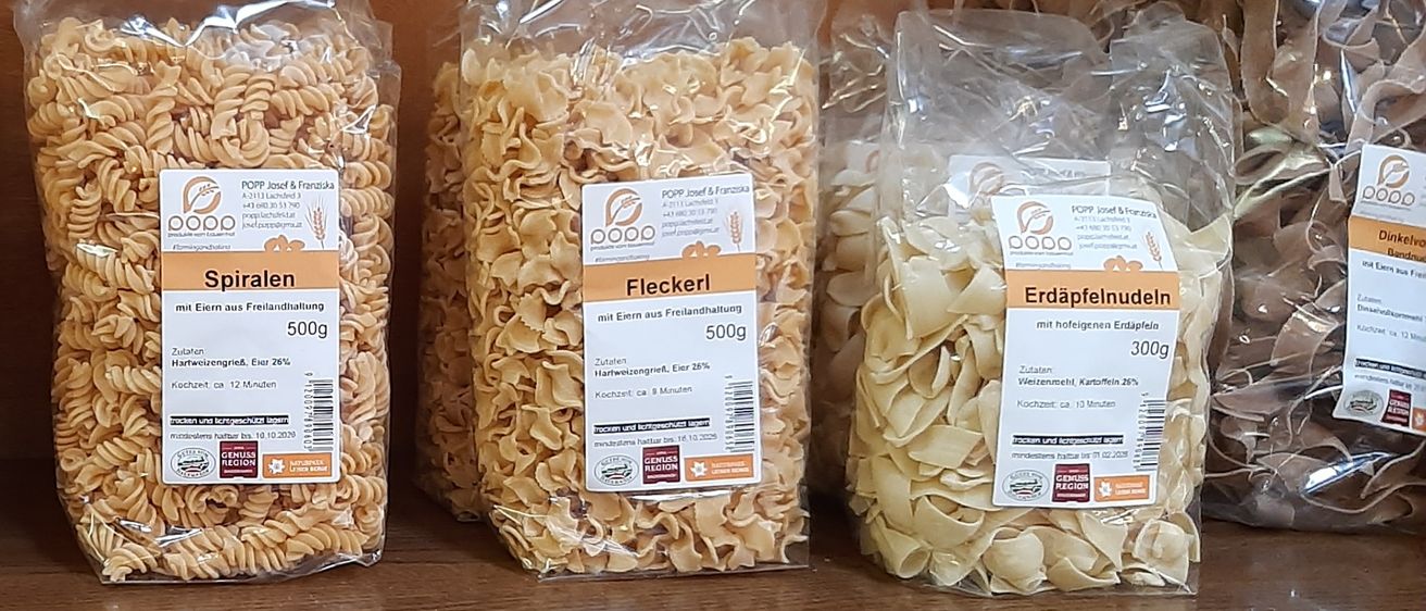 Ein Holzregal mit mehreren Tüten Pasta. Von links nach rechts: Bandnudeln, Spiralen, Elenkerl und Erdäpfelnudeln. Alle Tüten haben weiße Etiketten mit Text und Nährwertangaben.