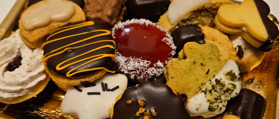 Ein goldfarbener Teller präsentiert eine Auswahl an schokoladenüberzogenen Keksen, darunter solche mit weißer Glasur, roter Füllung und Schokoladenspiralen. Andere Kekse haben Nüsse, Kokosnuss und dekorative Toppings.