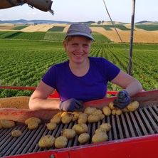Eine Frau in einem lila Shirt und Hut lächelt und hält Kartoffeln auf einer roten Maschine. Der Hintergrund zeigt ein weites grünes Feld und ein goldenes Feld.