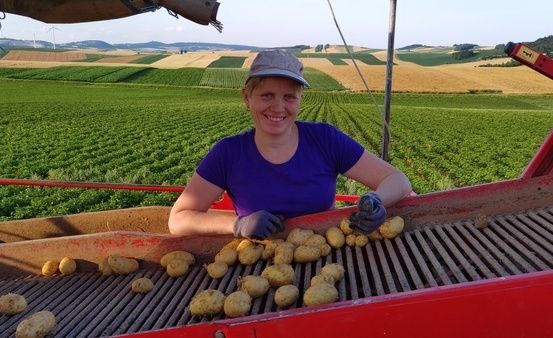 Eine Frau in einem lila Shirt und Hut lächelt und hält Kartoffeln auf einer roten Maschine. Der Hintergrund zeigt ein weites grünes Feld und ein goldenes Feld.