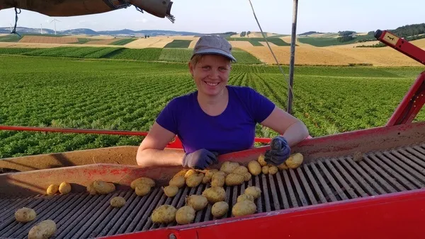 Eine Frau in einem lila Shirt und Hut lächelt und hält Kartoffeln auf einer roten Maschine. Der Hintergrund zeigt ein weites grünes Feld und ein goldenes Feld.