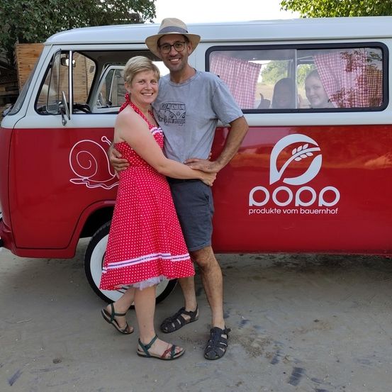 Ein Mann und eine Frau stehen gemeinsam vor einem rot-weißen Van mit einer Schnecke und einem Logo auf der Seite. Hinter ihnen sitzt ein Kind im Van.