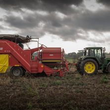 Eine rote Erntemaschine wird von einem grünen Traktor auf einem Feld unter einem stürmischen Himmel gezogen.