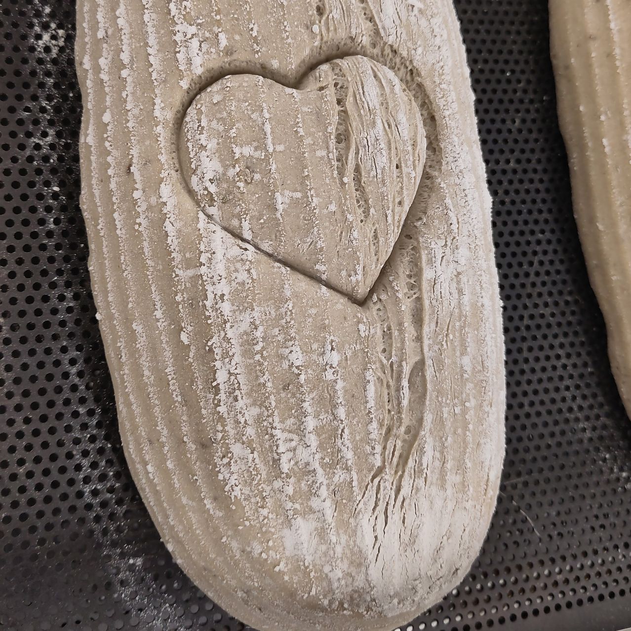 Ein herzförmiger Teig auf einer perforierten Oberfläche, möglicherweise einem Backstein, ist mit Mehl bestreut. Er hat eine leichte Textur mit einem rustikalen Look.