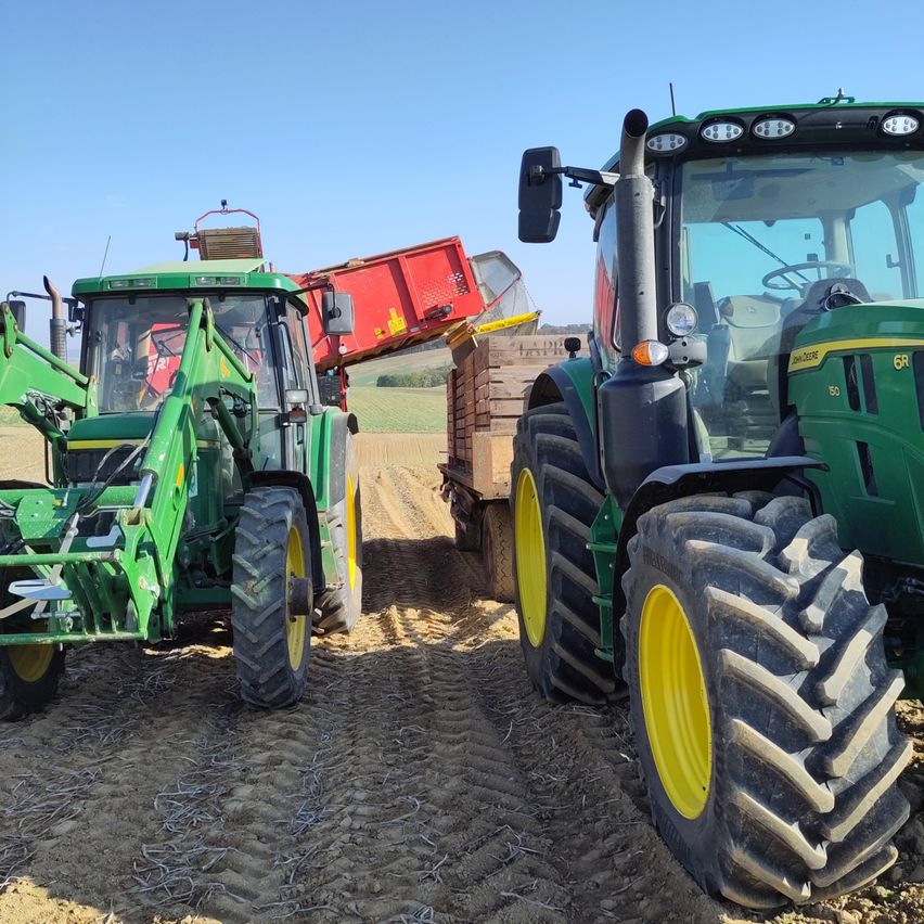 Zwei grüne Traktoren auf einem Bauernhof, einer entlädt einen roten Container in eine Holzkiste. Der Boden ist mit Erde und kleinem Gras bedeckt.