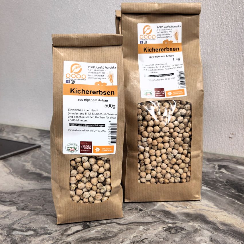 Zwei Packungen getrockneter Bohnen liegen auf einem Marmortresen. Jeder Beutel ist braun mit einem weißen Etikett. Die Etiketten enthalten den Namen 'Kichererbsen' und '500g'.