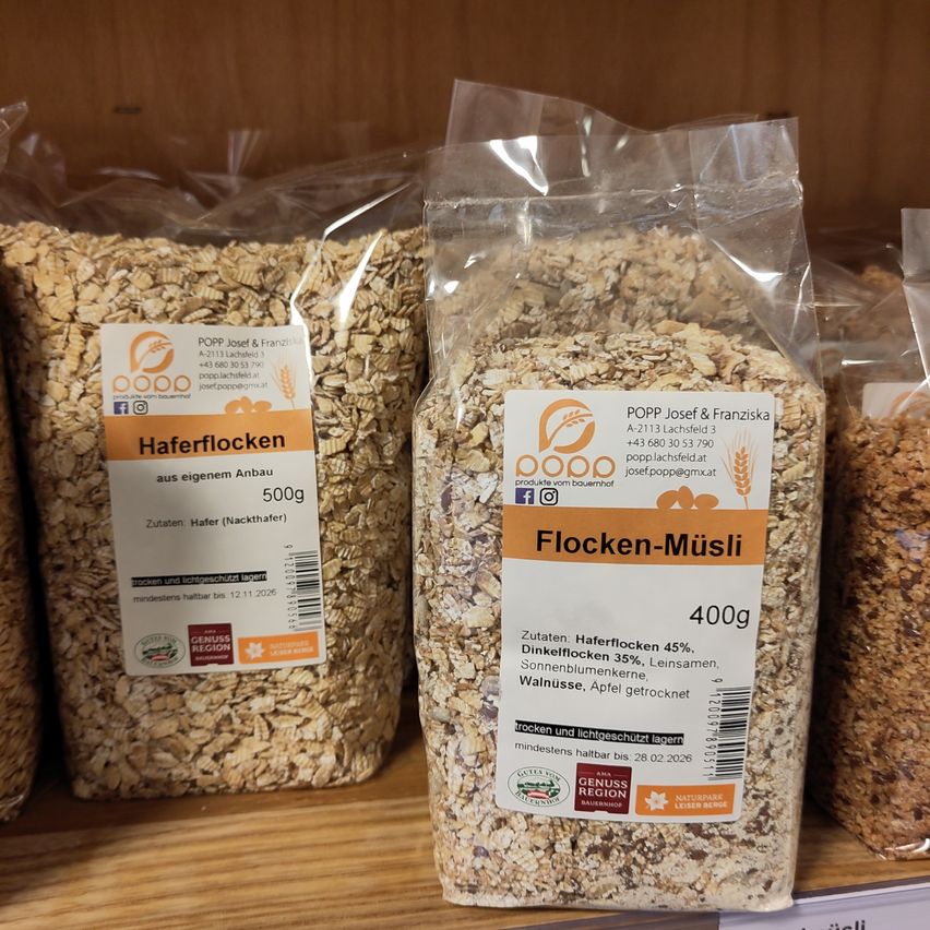 Zwei Beutel Haferflocken und Flocken-Muesli auf einem Holzregal. Der 'Haferflocken'-Beutel enthält 500g Haferflocken. Der 'Flocken-Muesli'-Beutel enthält 400g mit Zutaten wie Haferflocken, Sonnenblumenkernen und getrockneten Äpfeln.