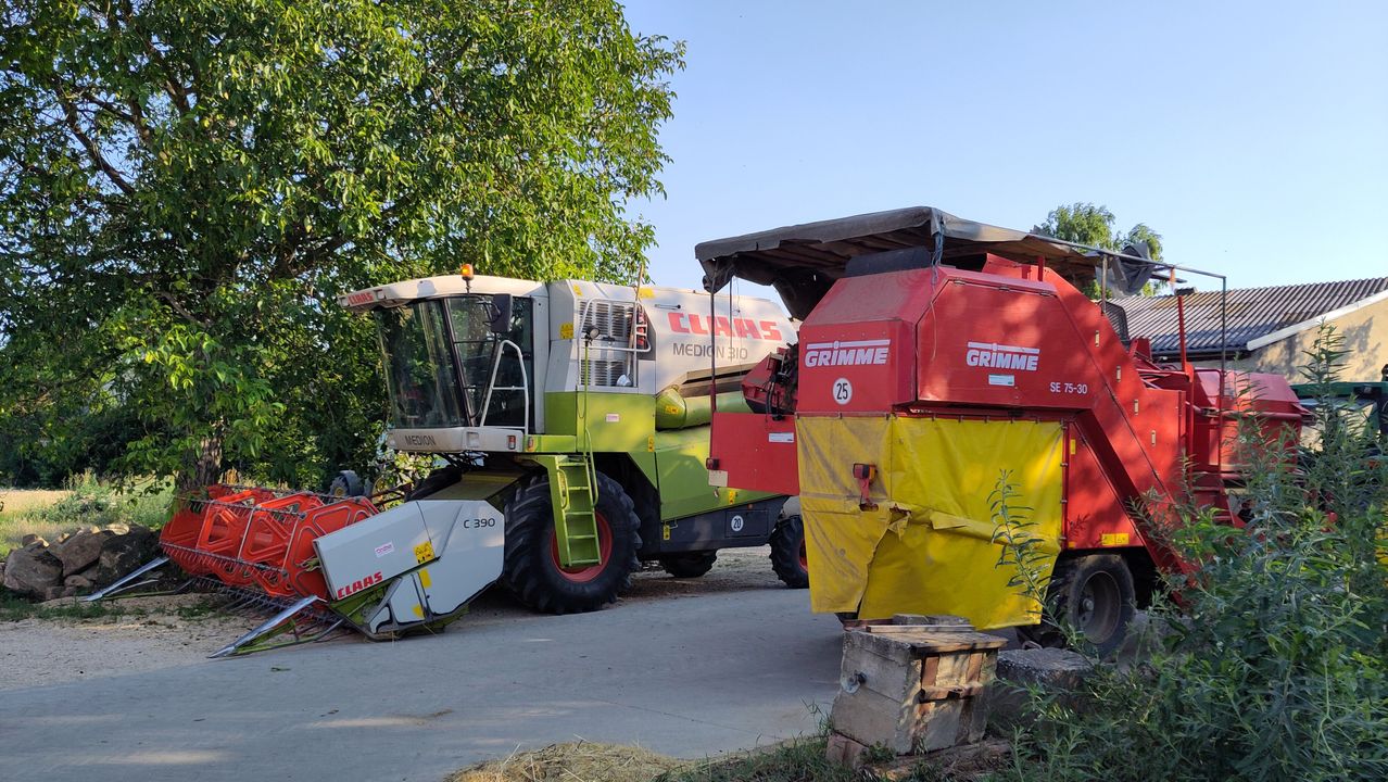 Zwei landwirtschaftliche Fahrzeuge, eines grün von Claas und das andere rot von Grimme, stehen unter einem Baum. Die Grimme hat eine beschädigte Plane und es fehlt ein Rad.