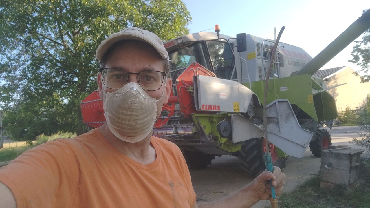 Ein Mann in einem orangefarbenen T-Shirt und einer Maske posiert mit einem CLAAS Traktor im Hintergrund. Er hält ein Werkzeug in der Hand.