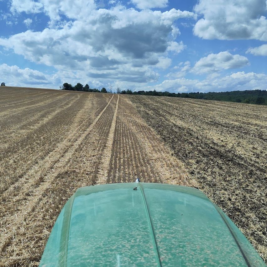 Ein Traktor fährt durch ein großes, trockenes Feld unter einem bewölkten blauen Himmel. Das Feld ist in Bereiche unterteilt, wobei einige Bereiche mehr Erde aufweisen als andere.