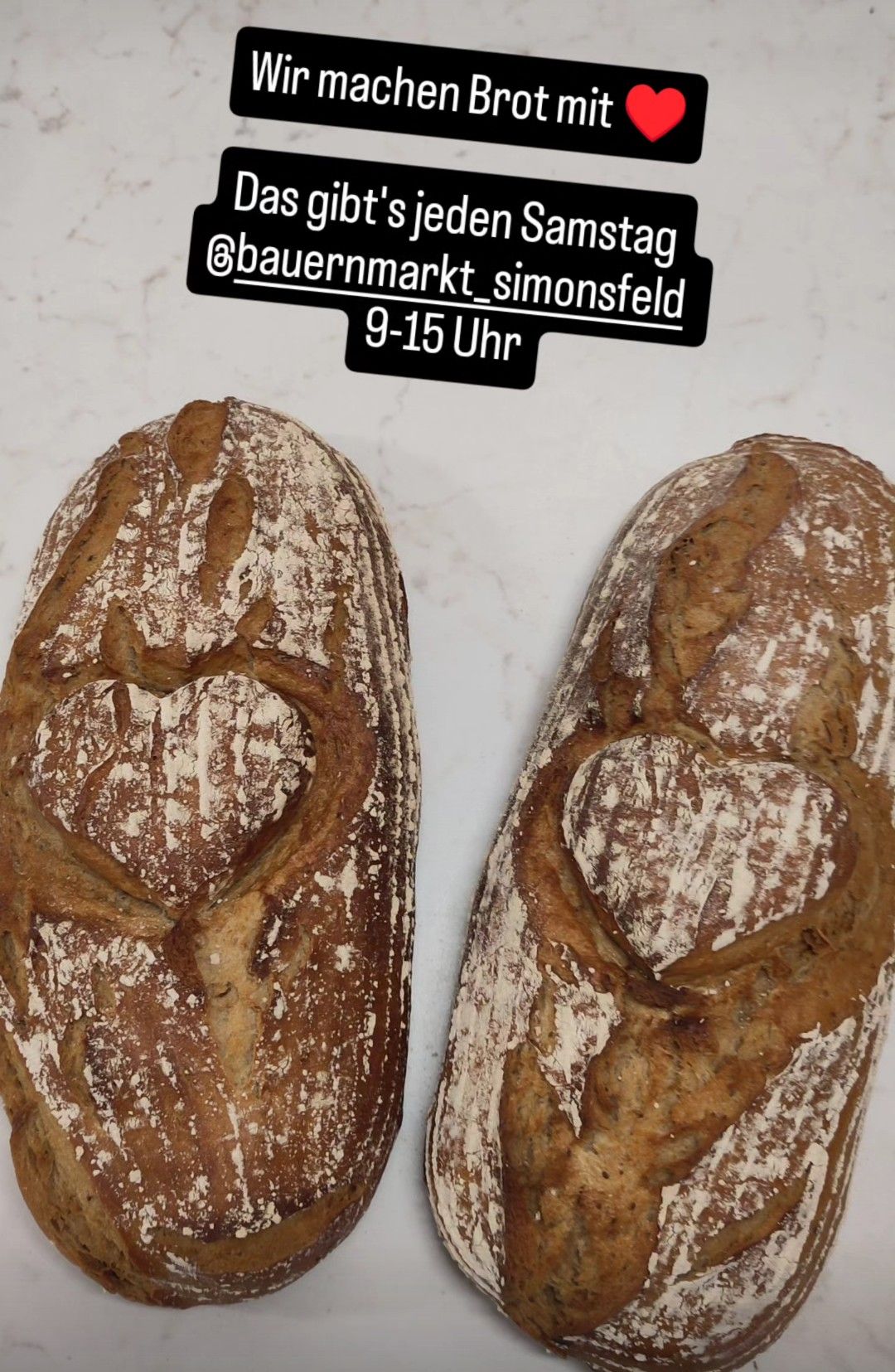 Zwei handwerklich geformte Brote in Herzform sind auf einer weißen Marmorfläche ausgestellt. Ein Etikett in der oberen Ecke lautet 'bauernmarkt_simonsfeld 9-15 Uhr'.