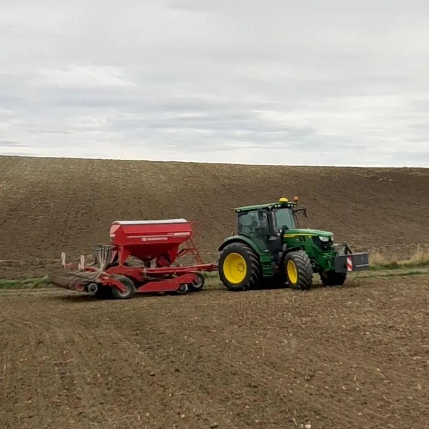 Ein grüner Traktor mit einem roten Sämaschinen arbeitet auf einem Feld mit einem Hügel im Hintergrund.
