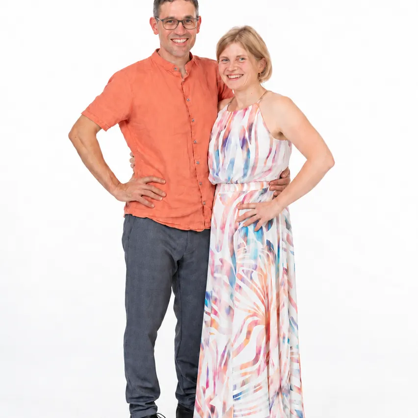 Ein Mann und eine Frau stehen gemeinsam im Studio, lächeln und posieren für ein Foto. Der Mann trägt ein orangefarbenes Hemd und graue Hosen, während die Frau ein farbenfrohes Kleid trägt.