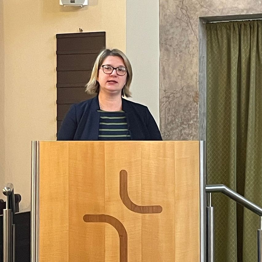 Eine Frau steht hinter einem Pult mit einem hölzernen Design, wahrscheinlich hält sie eine Rede. Sie trägt eine Brille und einen dunklen Pullover. Rechts befindet sich ein grüner Vorhang.