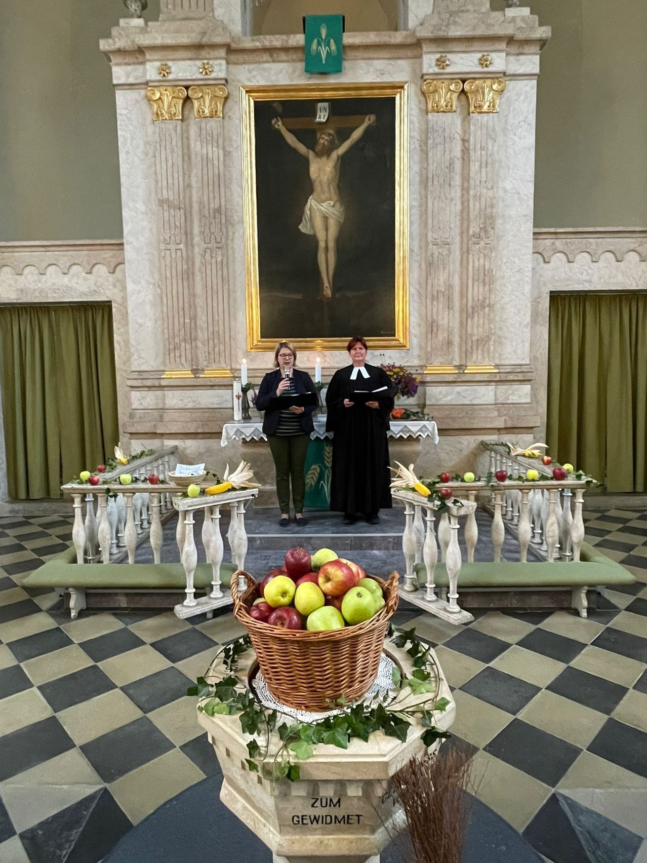 Zwei Frauen stehen in einer Kirche und halten Bücher. Ein Gemälde von Jesus am Kreuz steht hinter ihnen. Vor ihnen steht ein Korb mit Äpfeln.