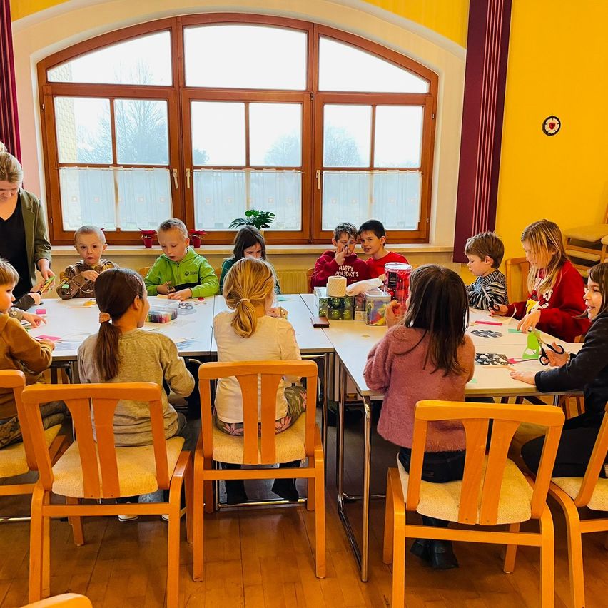 Kinder sitzen um Tische in einem Klassenzimmer und machen Bastelarbeiten. Eine Lehrerin beaufsichtigt sie in der Nähe eines Fensters mit Bäumen draußen.