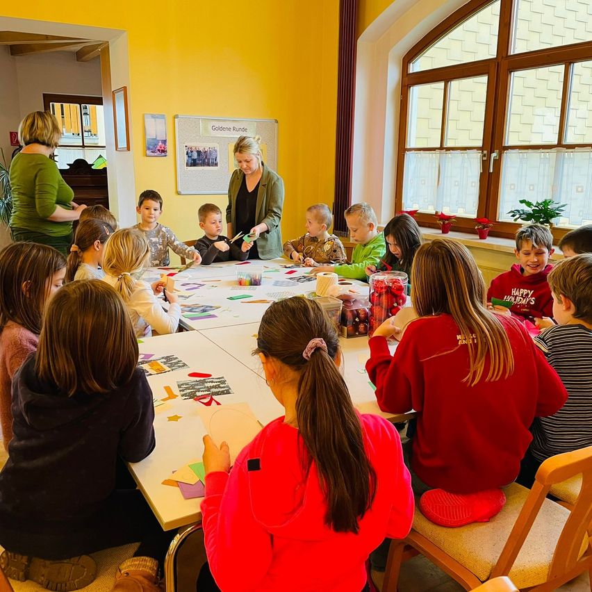 Ein Klassenzimmer mit gelben Wänden, Kinder sitzen um einen Tisch herum und machen Kunst, mit einer Frau, die vorne steht, einem Bulletin Board mit Bildern hinter ihr und einem Fenster mit Pflanzen.