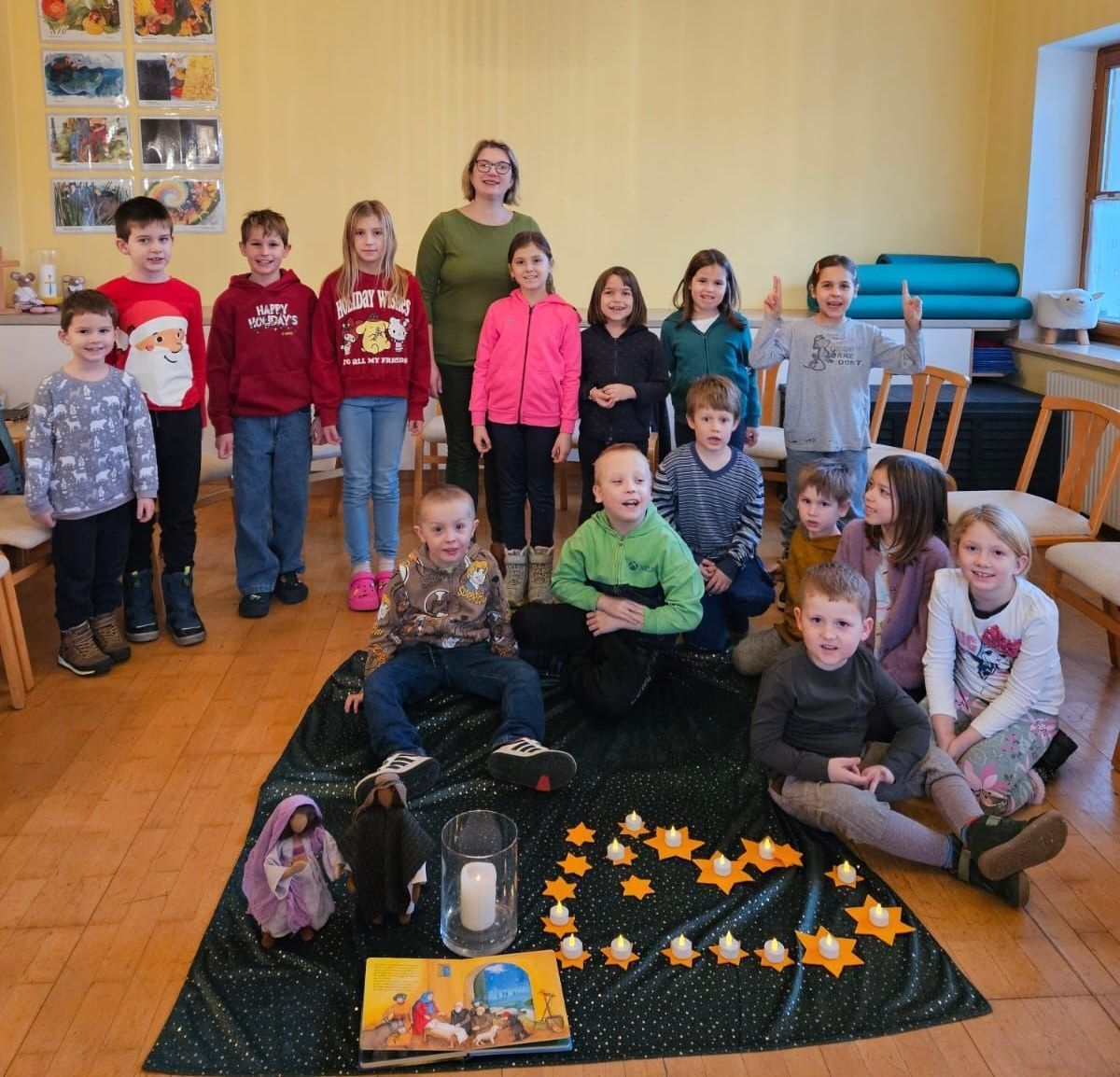 Eine Gruppe von Kindern und einer Frau versammelt sich in einem Raum. Sie lächeln und posieren für ein Foto. Eine Kerze ist auf einem Tisch mit einem Buch und Figuren angezündet. Der Raum hat Holzböden und gelbe Wände mit Bildern.