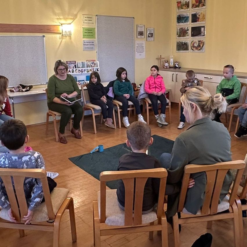 Eine Gruppe von Kindern und zwei Frauen sitzen in Stühlen in einem Klassenzimmer. Eine Frau liest ein Buch. Eine andere Frau, die neben einem Jungen sitzt, hält seine Schulter. Der Raum hat gelbe Wände und Holzböden.