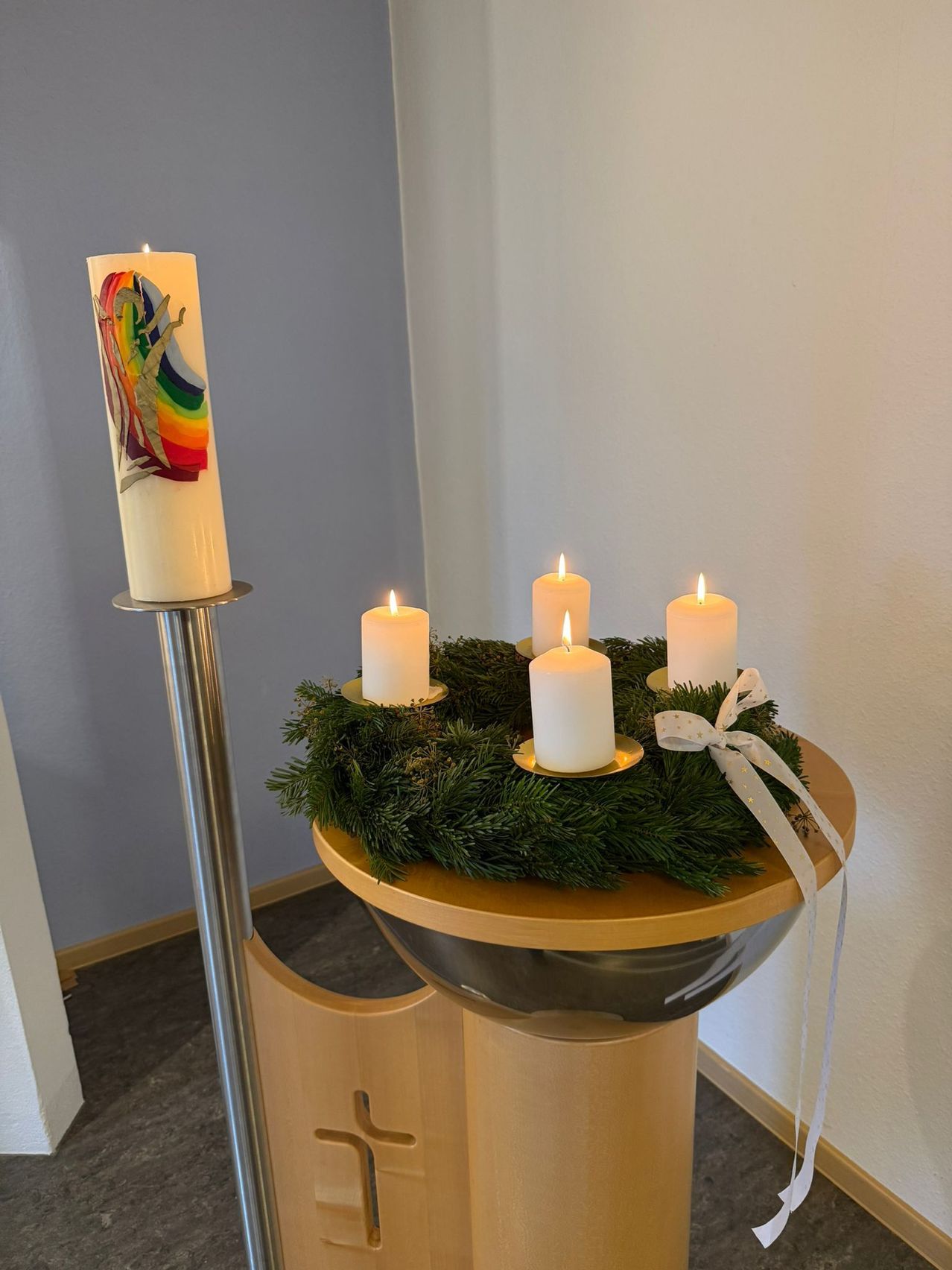 Ein Kerzenständer mit sechs Kerzen steht auf einem runden Holztisch, geschmückt mit einem Kranz aus grünen Tannenzweigen und einer weißen Schleife. Die Kerzen sind angezündet, und eine größere Kerze mit einem Regenbogendesign steht allein auf einem Metallkerzenhalter.