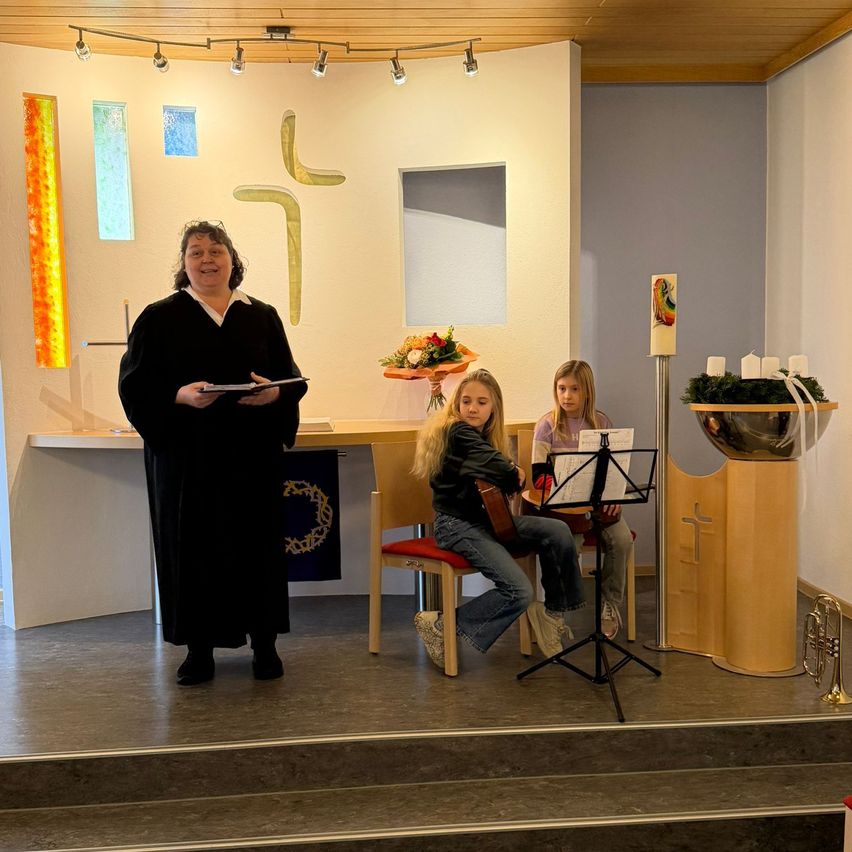 Eine Frau steht auf der Bühne und hält ein Buch in der Hand, möglicherweise leitet sie eine Zeremonie. In der Nähe sitzen zwei Mädchen, eines mit einer Gitarre, das andere mit Notenblättern. Hinter ihnen steht ein Tisch mit einer Blumenvase. Der Raum ist mit religiösen Symbolen und einer Trompete ausgestattet.