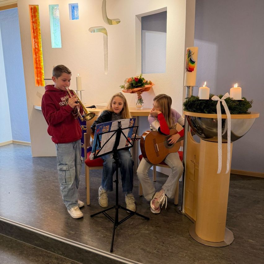 Drei Kinder spielen in einem Raum, ein Junge spielt Trompete und zwei Mädchen spielen Gitarre. Der Raum hat ein weihnachtliches Thema mit Kerzen und einer geschmückten Pflanze.
