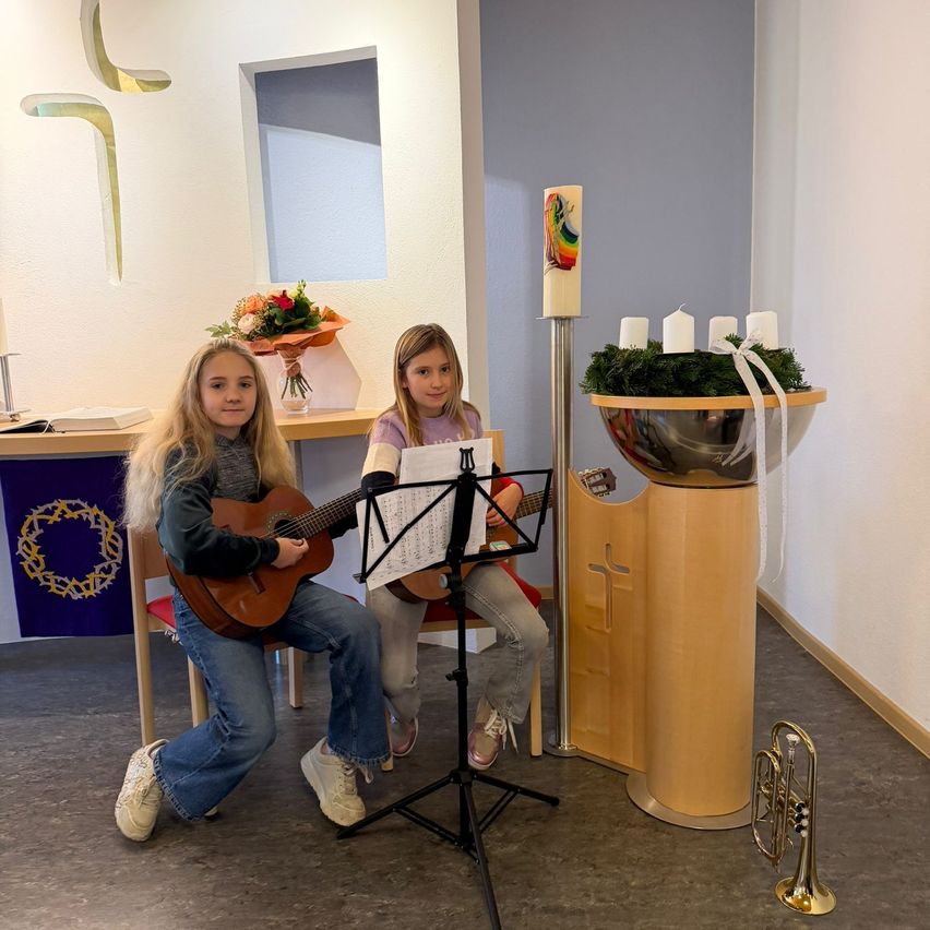 Zwei junge Mädchen sitzen auf Stühlen in einer Kirche und spielen Gitarre. In der Nähe steht eine Trompete. Vor ihnen befinden sich eine Blumenarrangement und Kerzen auf einem Ständer.