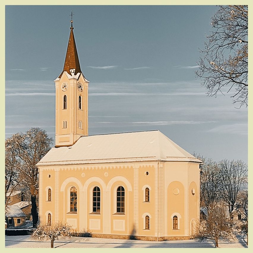 Eine Kirche mit hohem Turm und Uhr steht in einer verschneiten Landschaft. Das Gebäude ist beige mit Bogenfenstern und ist von kahlen Bäumen umgeben.