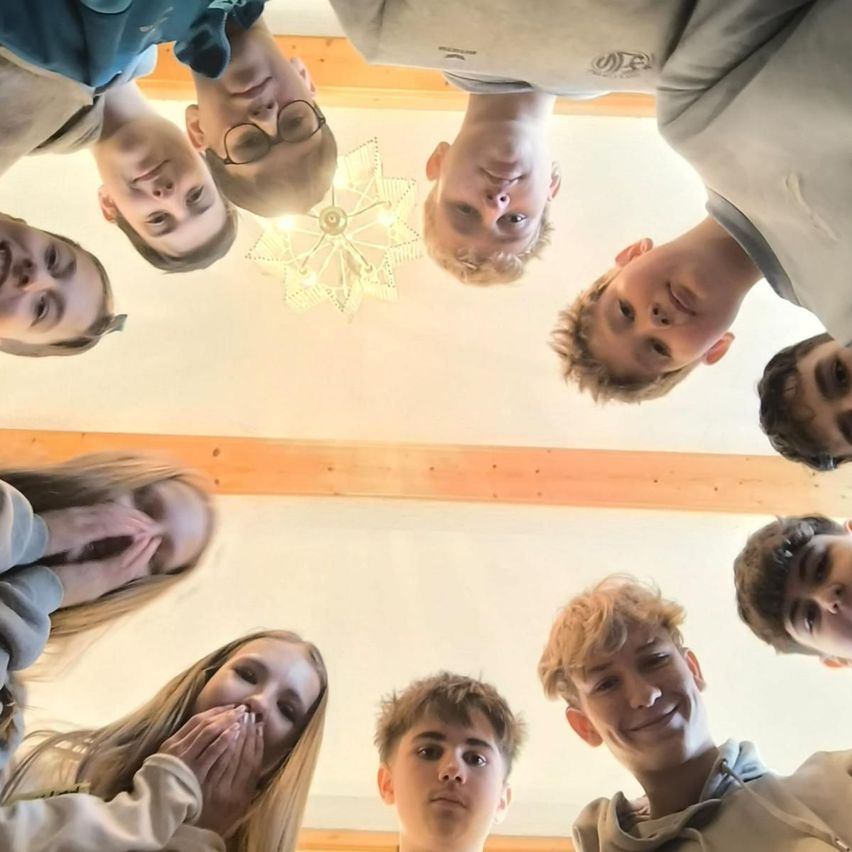 Eine Gruppe junger Menschen, darunter Jungen und Mädchen, sind versammelt und lächeln für ein Foto. Sie stehen in einer kreisförmigen Formation mit nach unten geneigtem Kopf. Eine Laterne hängt von der Decke über ihnen.