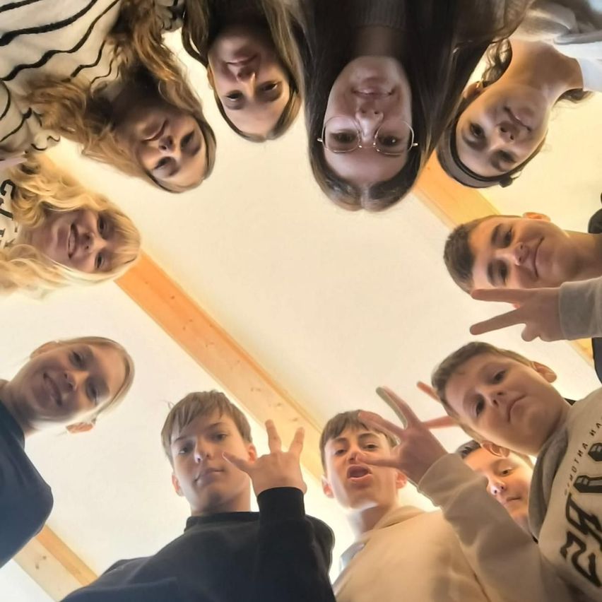 Eine Gruppe junger Menschen bildet einen Kreis, lächelt und macht Handzeichen. Sie scheinen für ein Foto in einem Raum mit Holzbalken zu posieren.