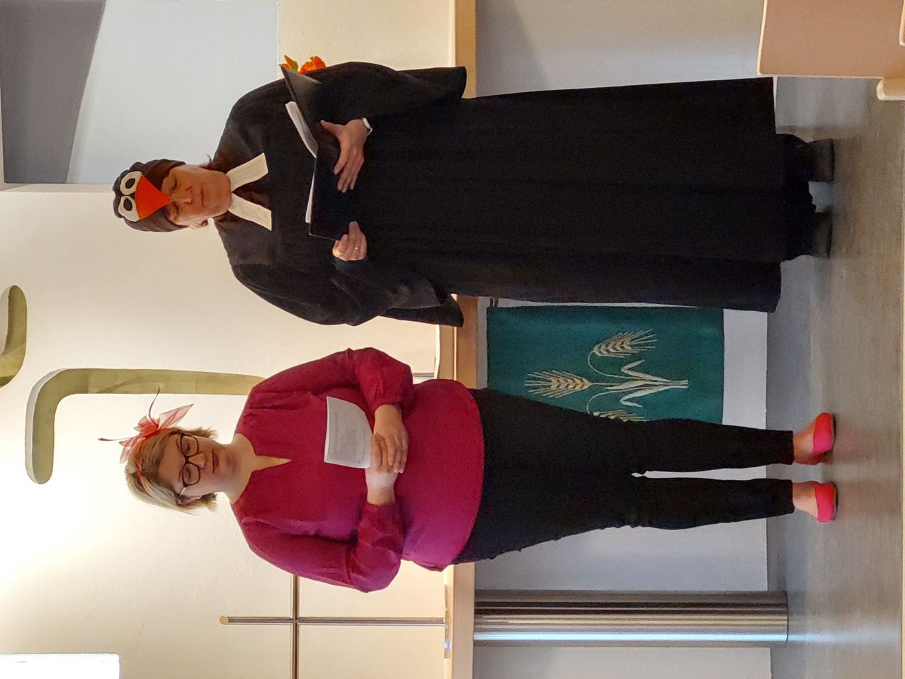 Zwei Frauen stehen in einem Raum. Die Frau vorn trägt ein rotes Oberteil und schwarze Hose. Die Frau dahinter trägt einen schwarzen Umhang und eine Vogel-Mütze. Beide halten Papiere. Ein grüner Stoff mit Weizenmustern hängt an der Wand hinter ihnen.