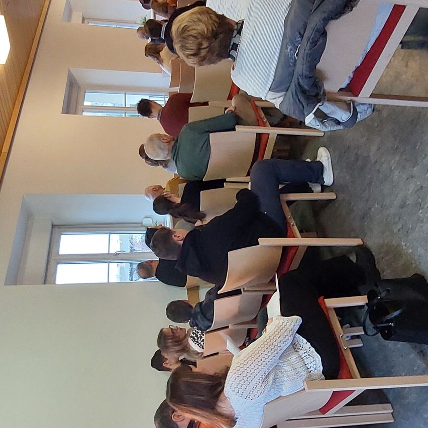 Eine Gruppe von Menschen sitzt in einem Klassenzimmer auf Stühlen. Einige tragen Brillen und andere haben den Kopf gesenkt. Eine Person in der Vorderreihe lehnt sich in ihrem Stuhl zurück.