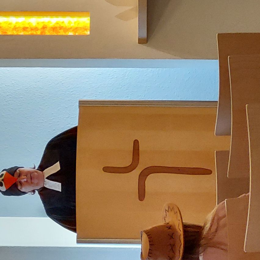 Eine Person in Kostüm steht hinter einem Podium mit Kreuzsymbol. Eine Person mit Hut sitzt vor einer Pappwand.