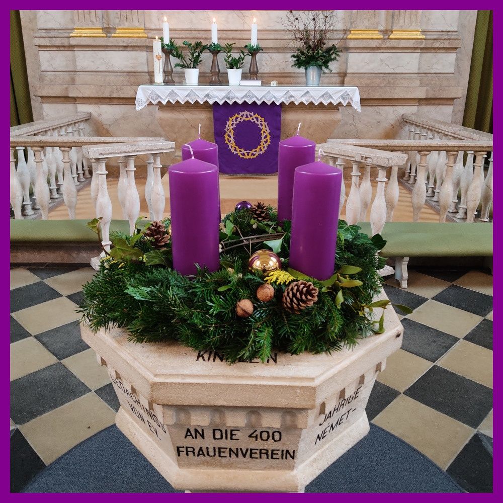 Eine kreisförmige Anordnung von lila Kerzen und Grünpflanzen auf einem Podest mit einem Kranz aus Tannenzapfen, Stechpalmenblättern und goldenen Ornamenten. Dahinter befindet sich ein Kirchenaltar mit weiteren Kerzen und Topfpflanzen.