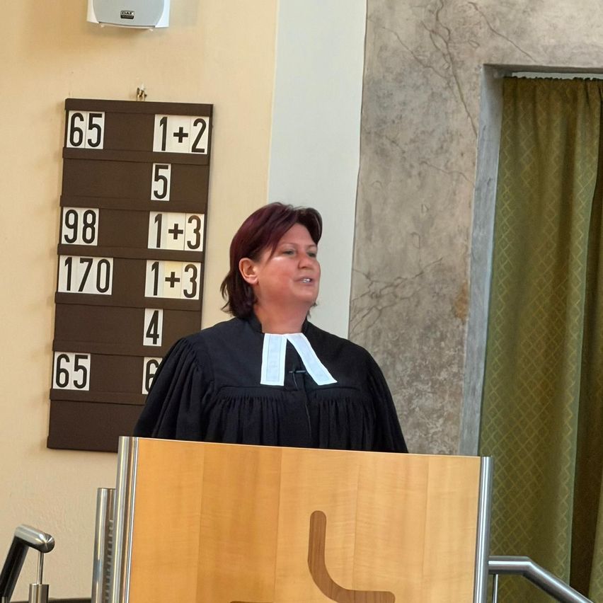 A woman wearing a black robe stands at a podium with a wooden sign. Behind her is a wall with a scoreboard displaying numbers 65, 98, 170, 4, and 65. There is also a microphone on the podium.