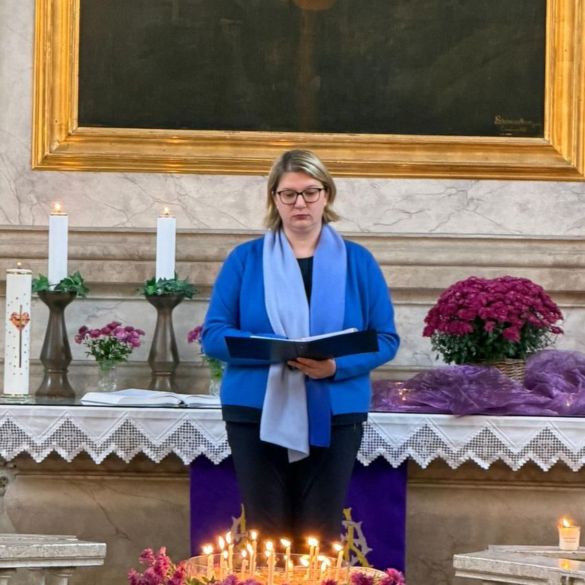A woman stands in front of an altar holding a book, wearing glasses and a scarf, with candles and flowers around her. Behind her is a painting on the wall.