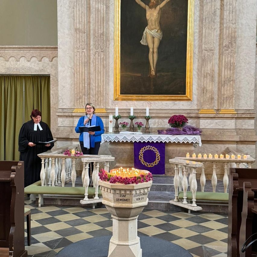 Two women stand in a church, one reading a book and the other speaking into a microphone. A cross is on the wall, and candles are lit on the altar.
