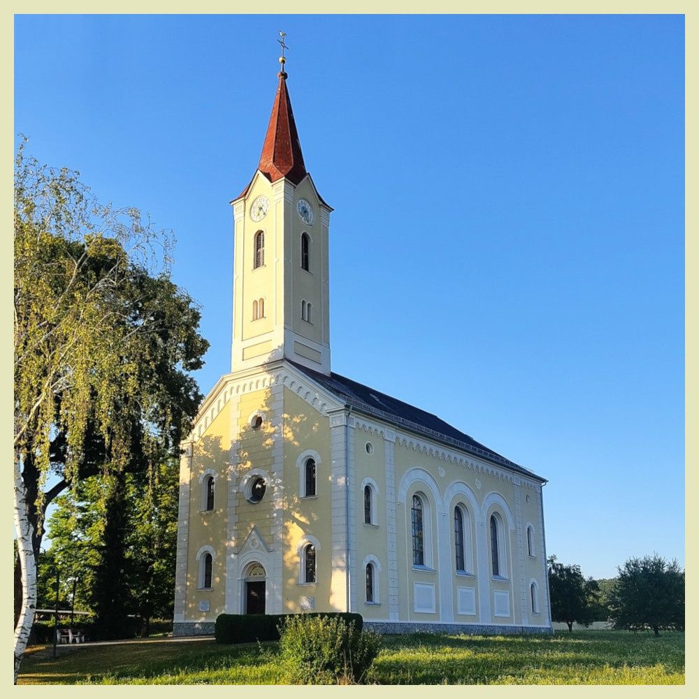 A small church with a steeple and a clock stands on a grassy field. The building is painted yellow with white accents. There are trees around it.