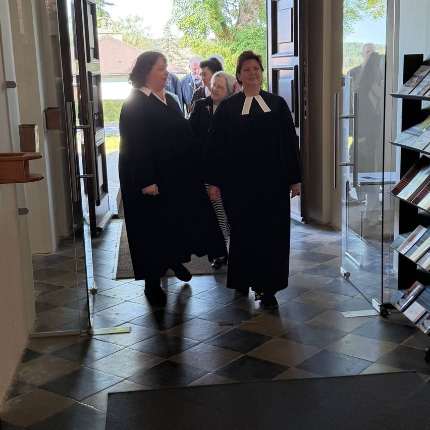 Two women in black robes walk through a door. Behind them, two people are standing near the open door. A shelf with books is to the right.