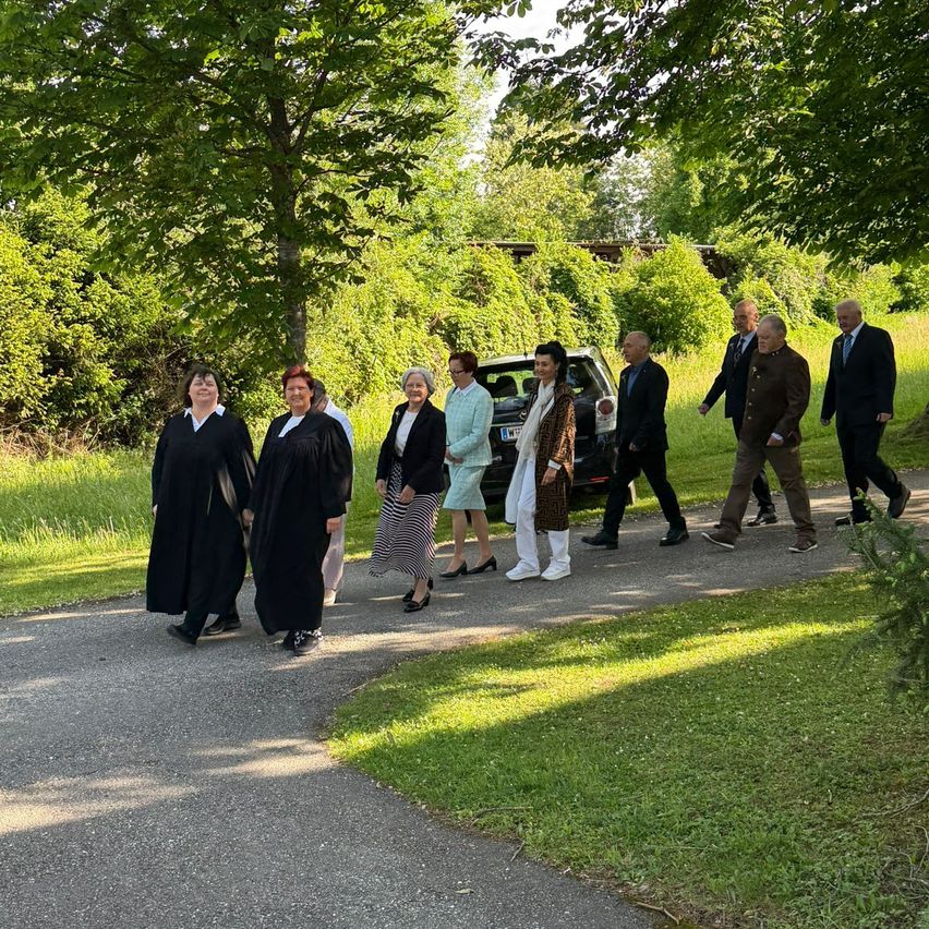 A group of people in formal attire walks on a pathway in a park, with trees and grass on both sides. In the background, a car is parked near a fence.