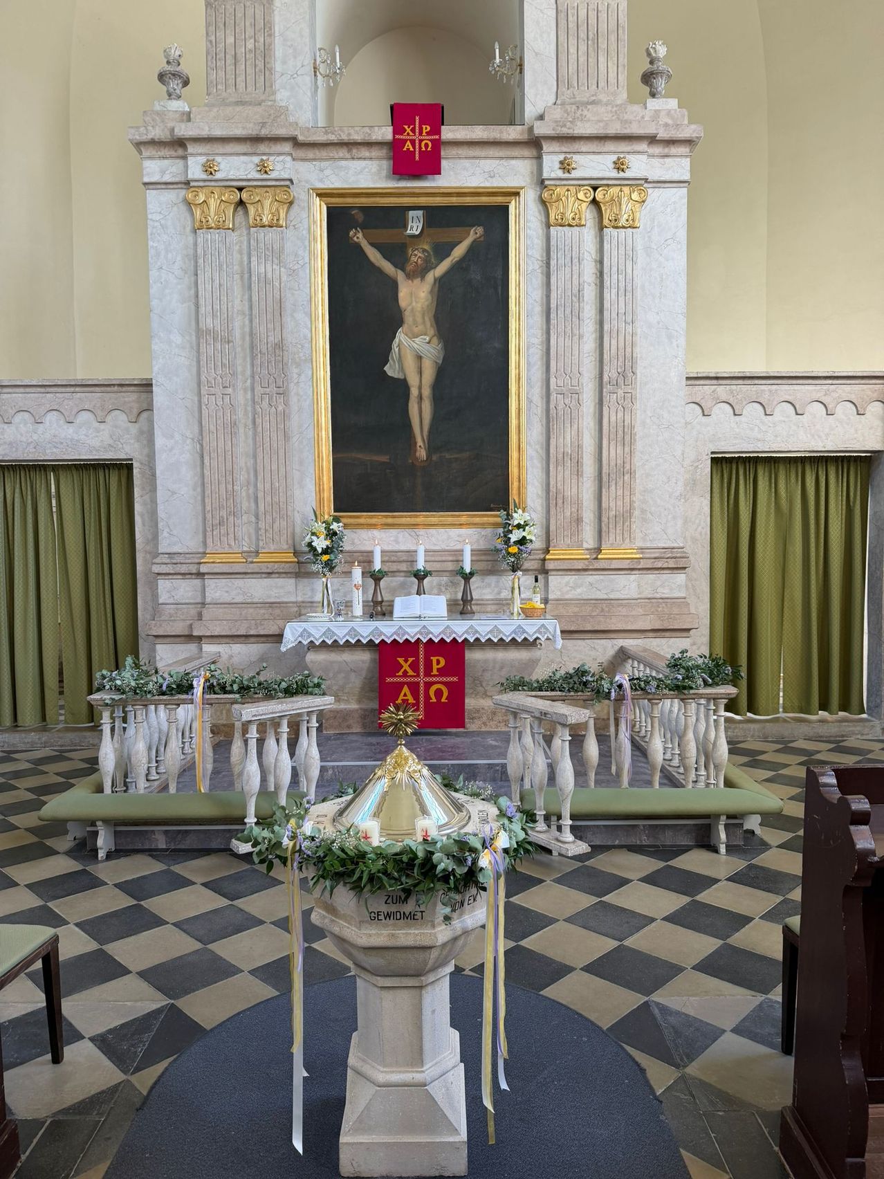 An altar adorned with a large crucifix, candles, and fresh flowers stands in a church sanctuary. The altar is flanked by two benches, and there is a checkered floor and green curtains.