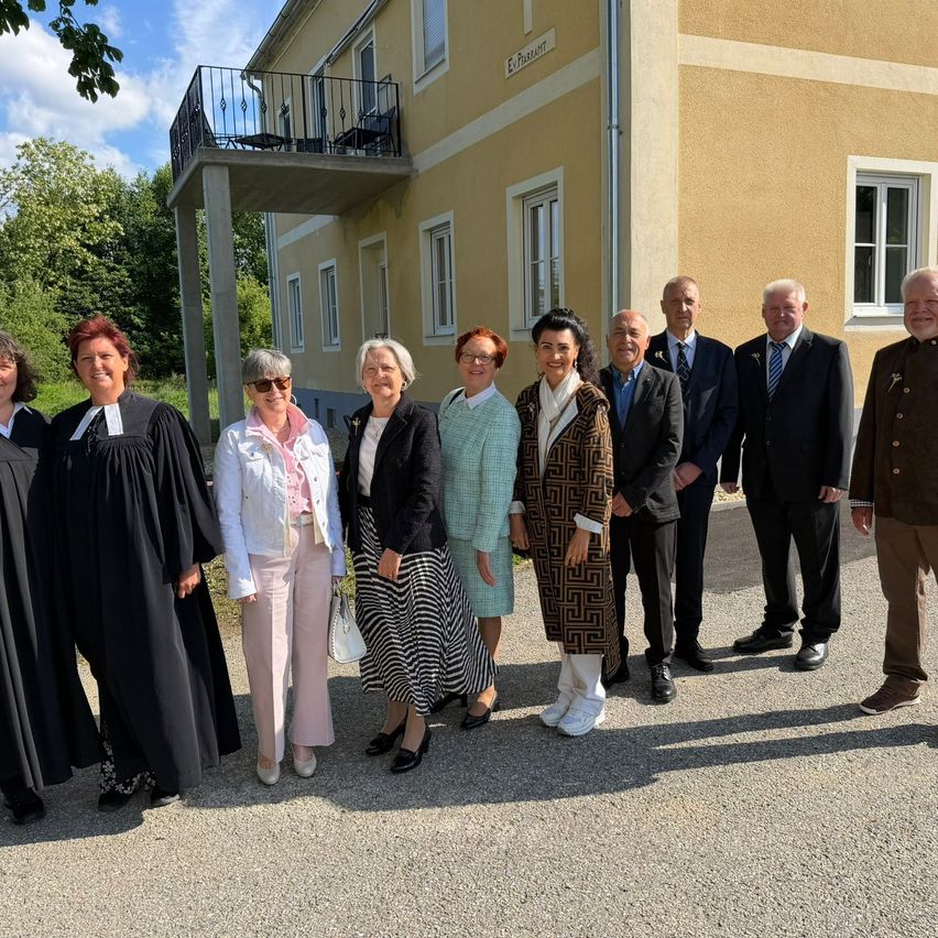 A group of people dressed in formal attire stand outside a building with a balcony. They are likely attending a formal event or meeting.
