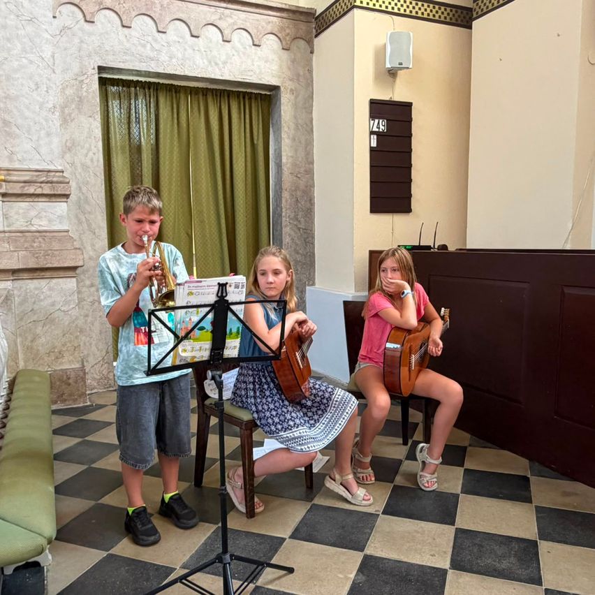 Drei Kinder spielen in einem Raum Musikinstrumente. Ein Junge spielt Trompete, während zwei Mädchen Gitarre spielen. Sie sitzen auf Stühlen mit einem Notenständer vor ihnen. Der Raum hat einen karierten Boden und eine Wand mit einem Vorhang und einem Lautsprecher.
