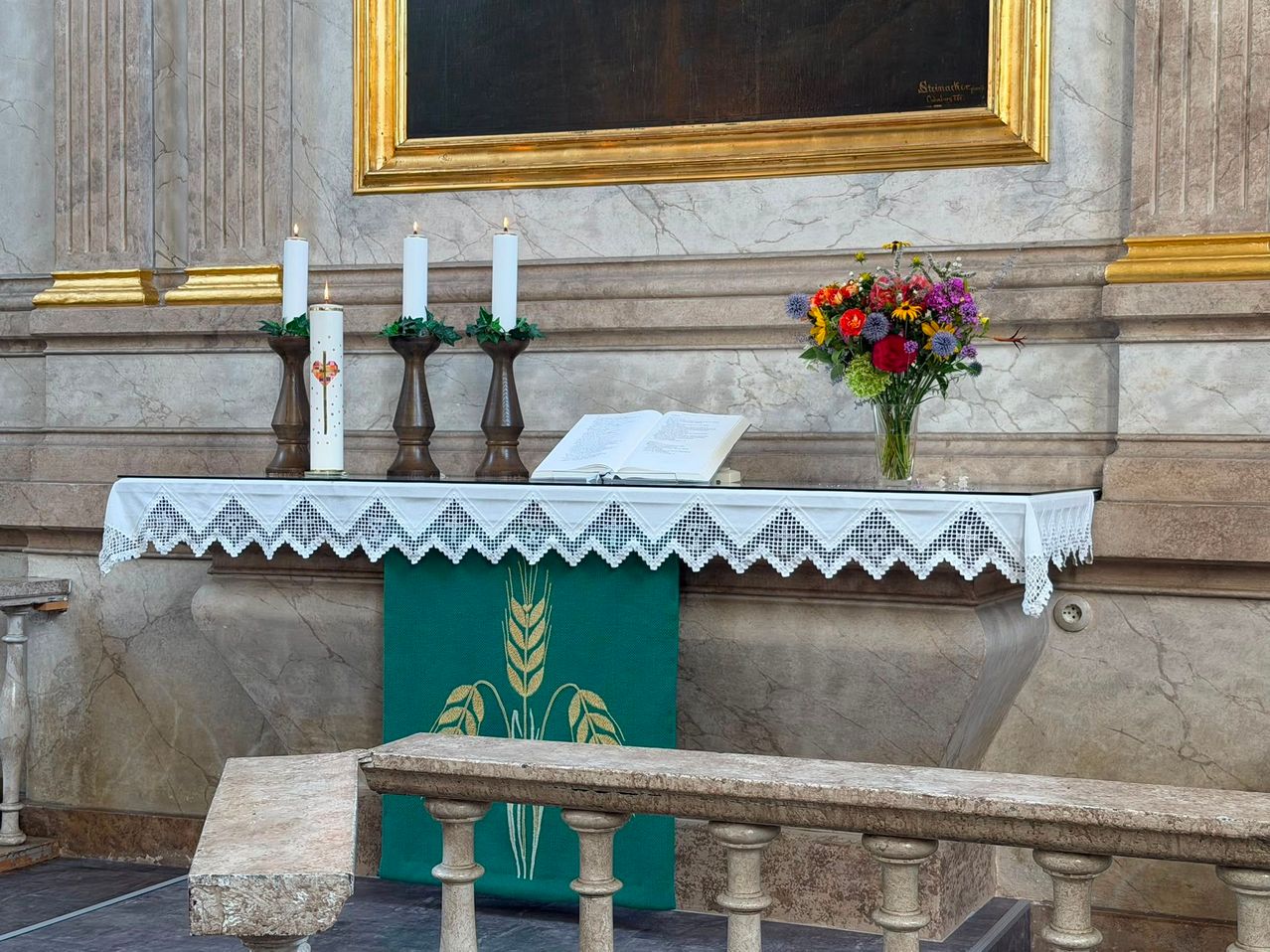 Ein Kirchenaltar mit drei Kerzen, einem Buch und einer Vase mit Blumen. Der Altar ist mit einem weißen Spitzentuch bedeckt und hat einen grünen Stoff mit Weizendesign. Ein goldgerahmtes Gemälde ist an der Wand darüber montiert.