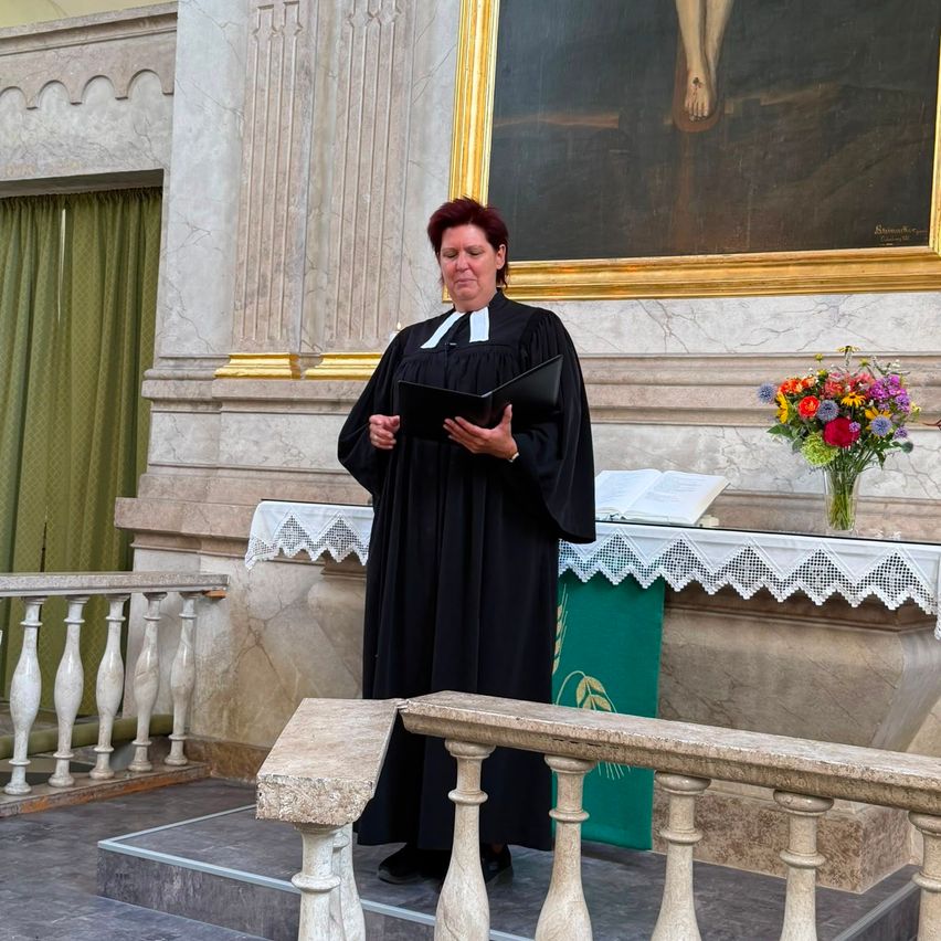 Eine Frau in einem schwarzen Gewand steht an einem Pult in einer Kirche, hält ein Buch in der Hand und schaut nach unten. Hinter ihr befindet sich ein großes Gemälde von Jesus am Kreuz.