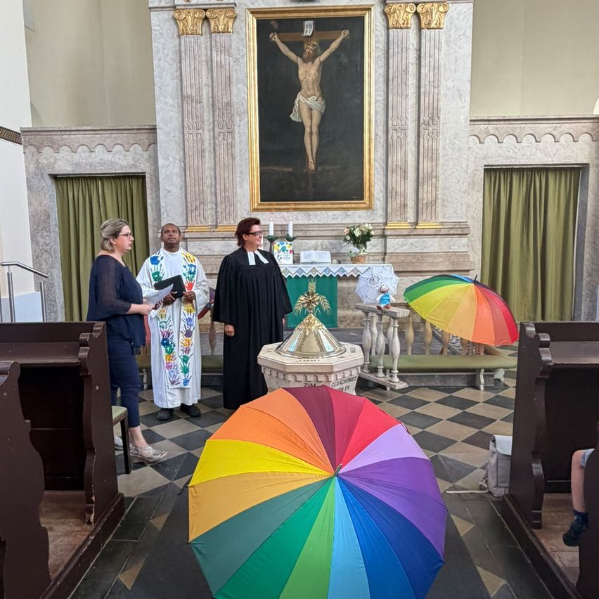 Drei Personen stehen in einer Kirche, eine hält einen bunten Regenschirm, eine andere liest aus einem Buch und eine dritte trägt einen schwarzen Umhang. Ein großes Gemälde von Jesus am Kreuz schmückt die Wand.
